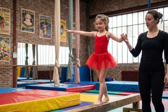 Jeune fille en justaucorps rouge apprenant l'équilibre sur une poutre dans une école de cirque à Paris avec une instructrice
