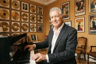 Luc Plamondon, compositeur québécois célèbre, pensif au piano dans un salon rempli de disques d'or et d'affiches de spectacles